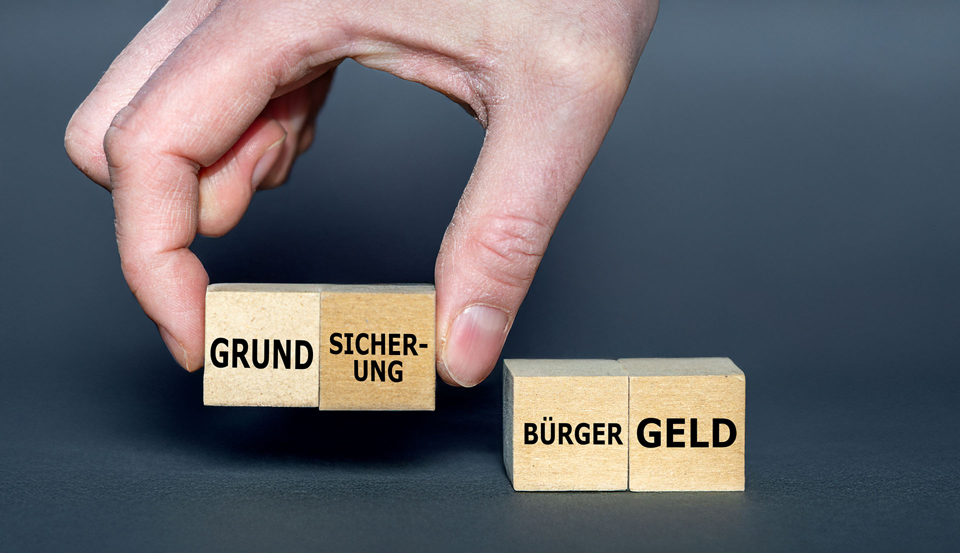 Ein Bild mit einer Hand und vier kleinen Holzwürfeln, die beschriftet sind mit den Begriffen: GRUND, SICHERUNG; BÜRGER und GELD. Die Hand greift mit zwei Fingern die Würfel mit dem Text: GRUNDSICHERUNG