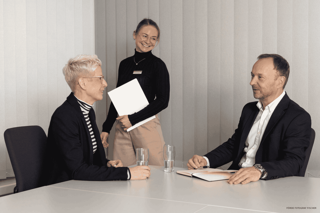 Zwei Menschen unterhalten sich an einem Tisch, ein weiterer Mensch läuft vorbei
